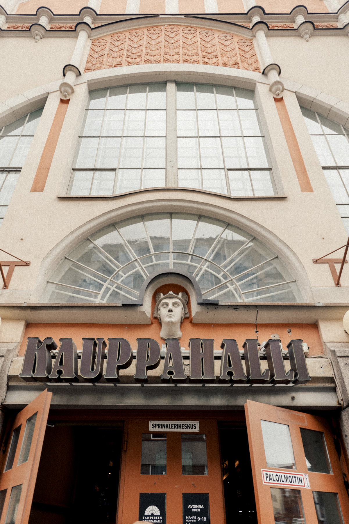 Entrance to a building labeled "Kauppahalli," with large windows and architectural details above the door, including a sculpture of a face.