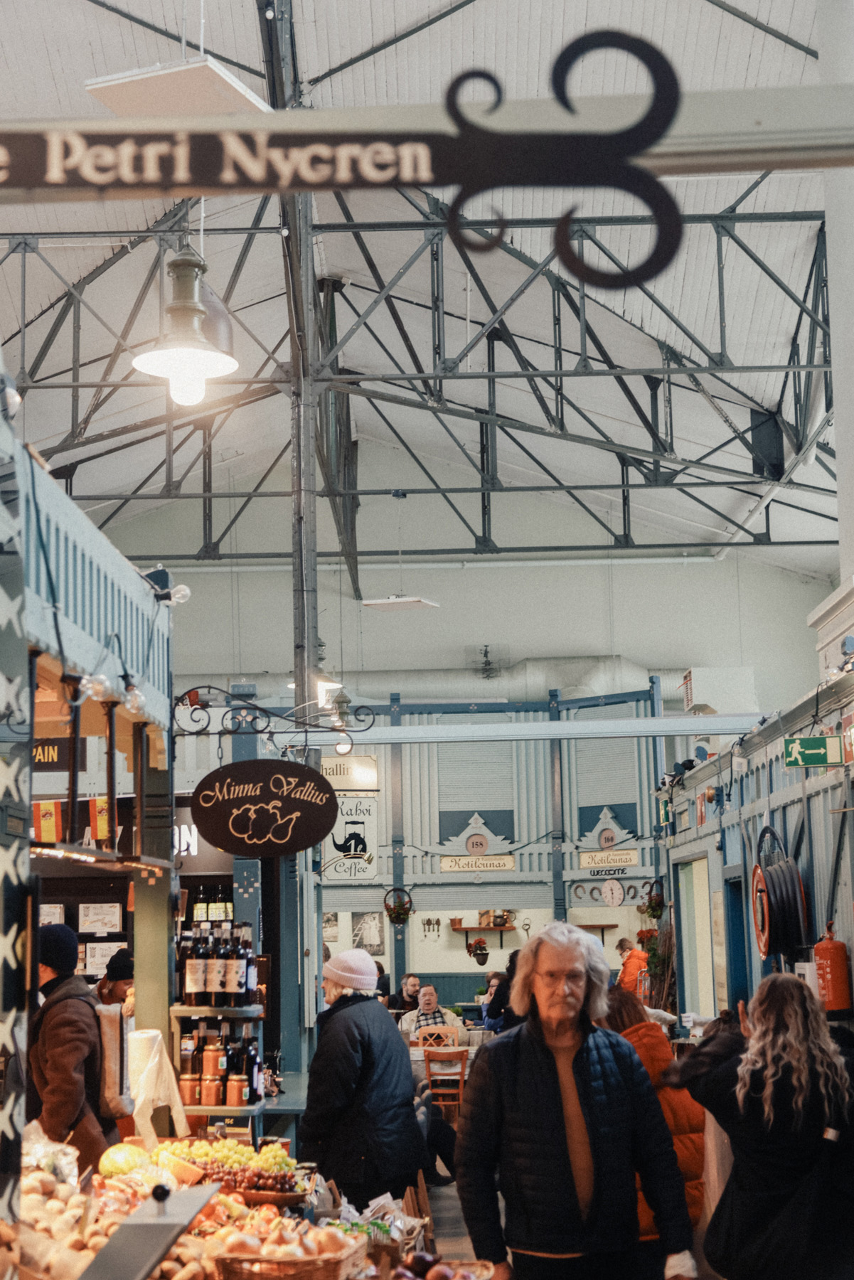 Indoor market scene with people shopping for goods at various stalls. A sign reading "Petri Nygren" and hanging scissors are prominent in the foreground.