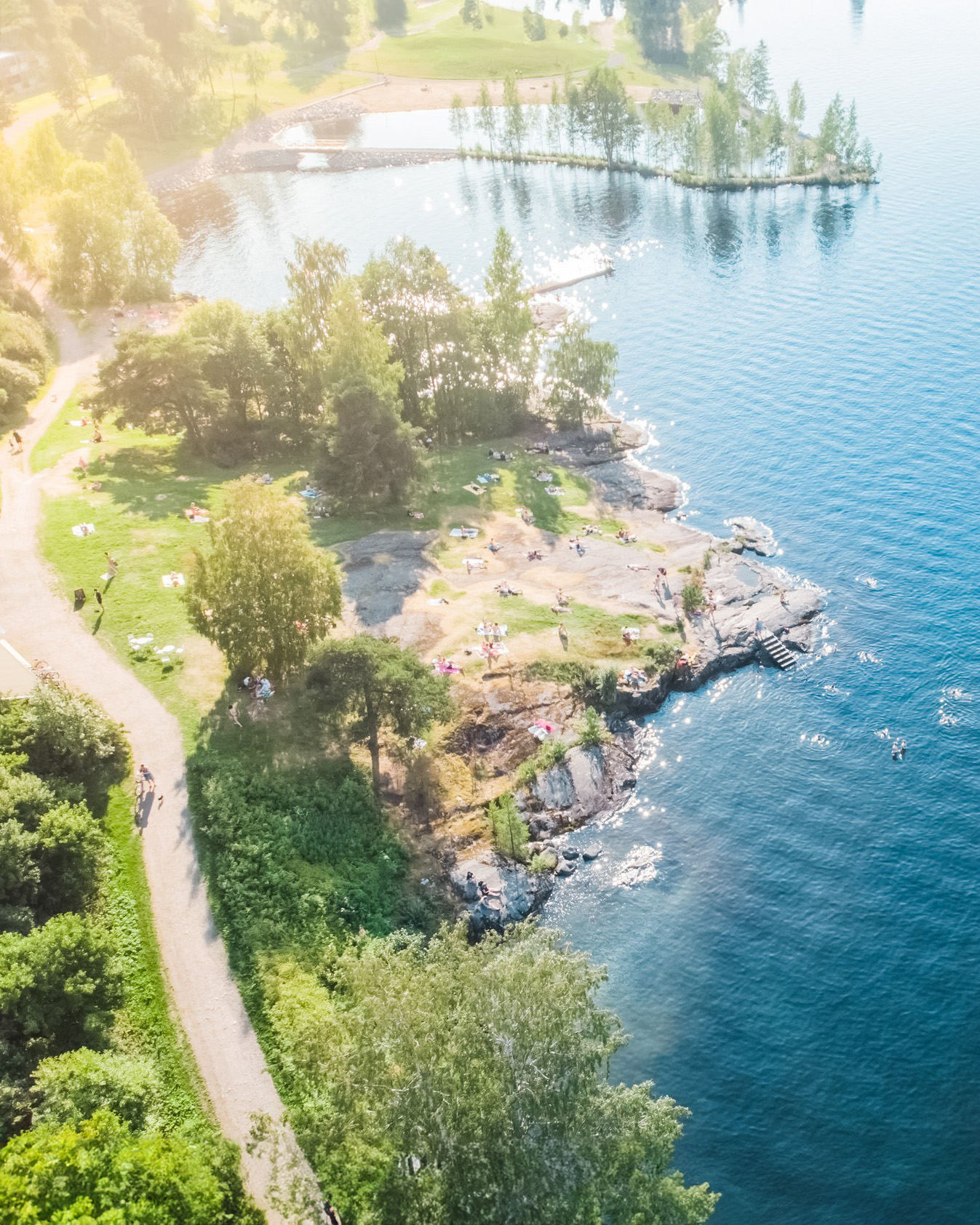 Aerial view of Eliander Beach with people relaxing on a grassy area, trees nearby, and a gravel path along the shore; the calm waters of Näsijärvi in Tampere, Finland, have a few swimmers enjoying the day.