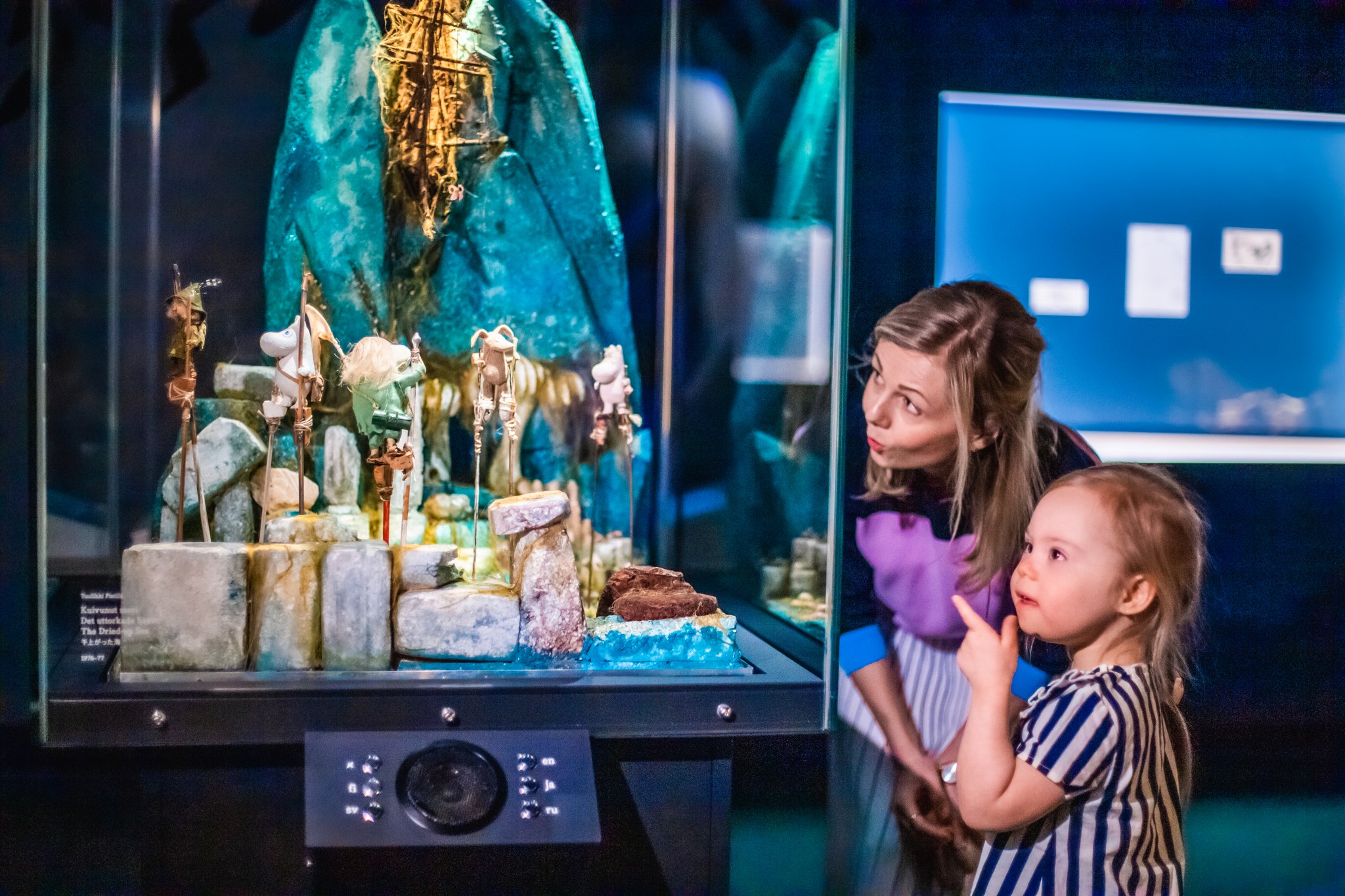 A woman and a child observe a detailed diorama in a glass display case, depicting a rocky landscape with miniature figures, at the Moomin Museum Tampere—a perfect spot for family fun.