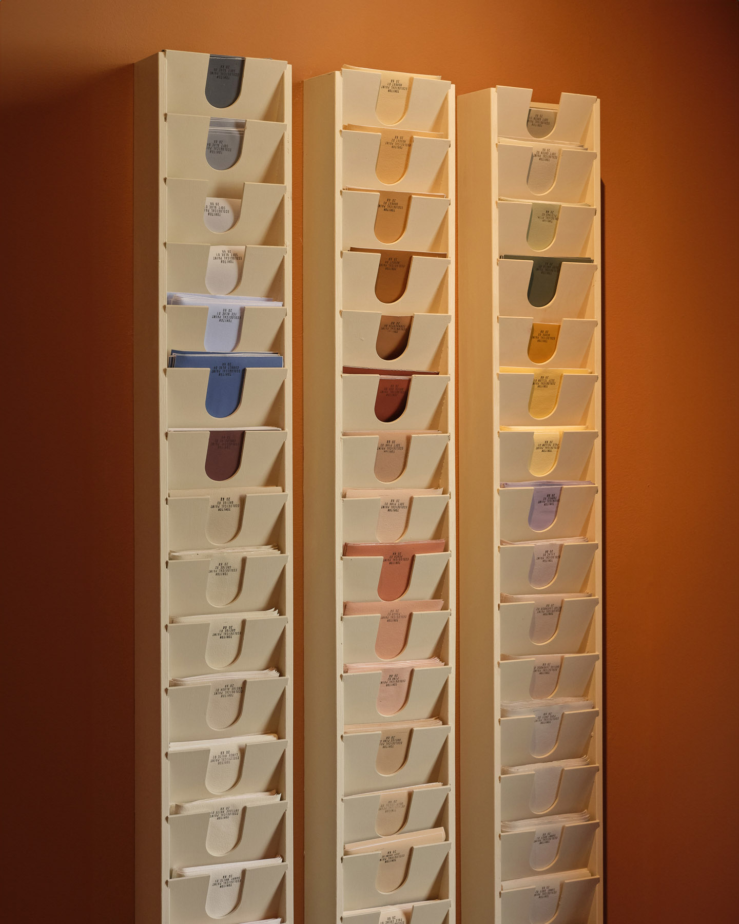 Three vertical racks of Toniton Wall Paint Samples are mounted on a wall in Boutique Stockholm, displaying various shades of neutral and warm colors. The background wall is painted in a warm terracotta hue.