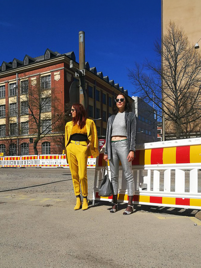 Two people stand in front of the Yesterday Shop in Tampere, framed by building and construction barriers under a clear blue sky. One person wears a yellow outfit; the other dons gray and holds a white bag.