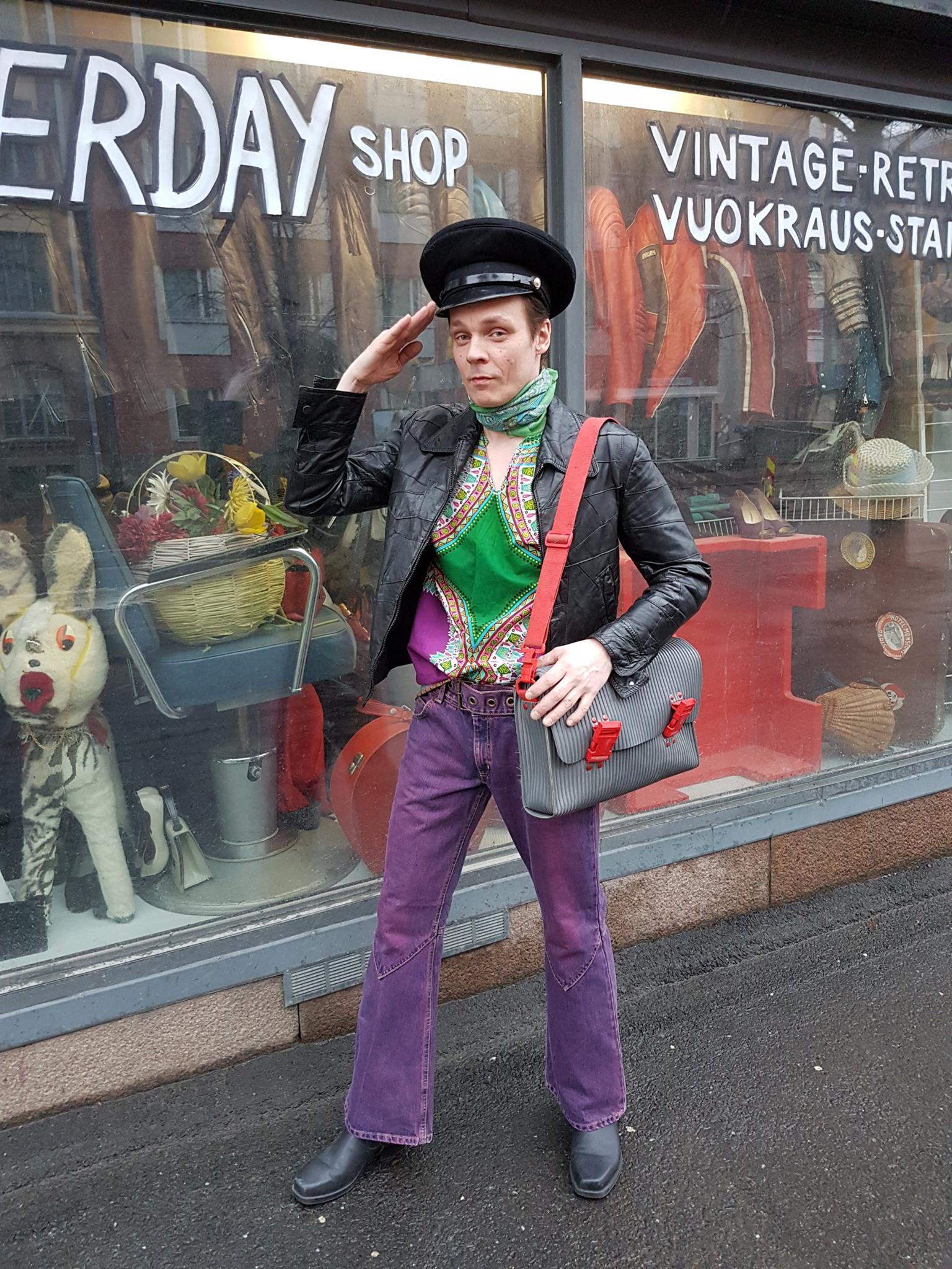 A person in a colorful outfit and black hat salutes in front of the Yesterday Shop Tampere window, displaying assorted retro items and a mannequin.