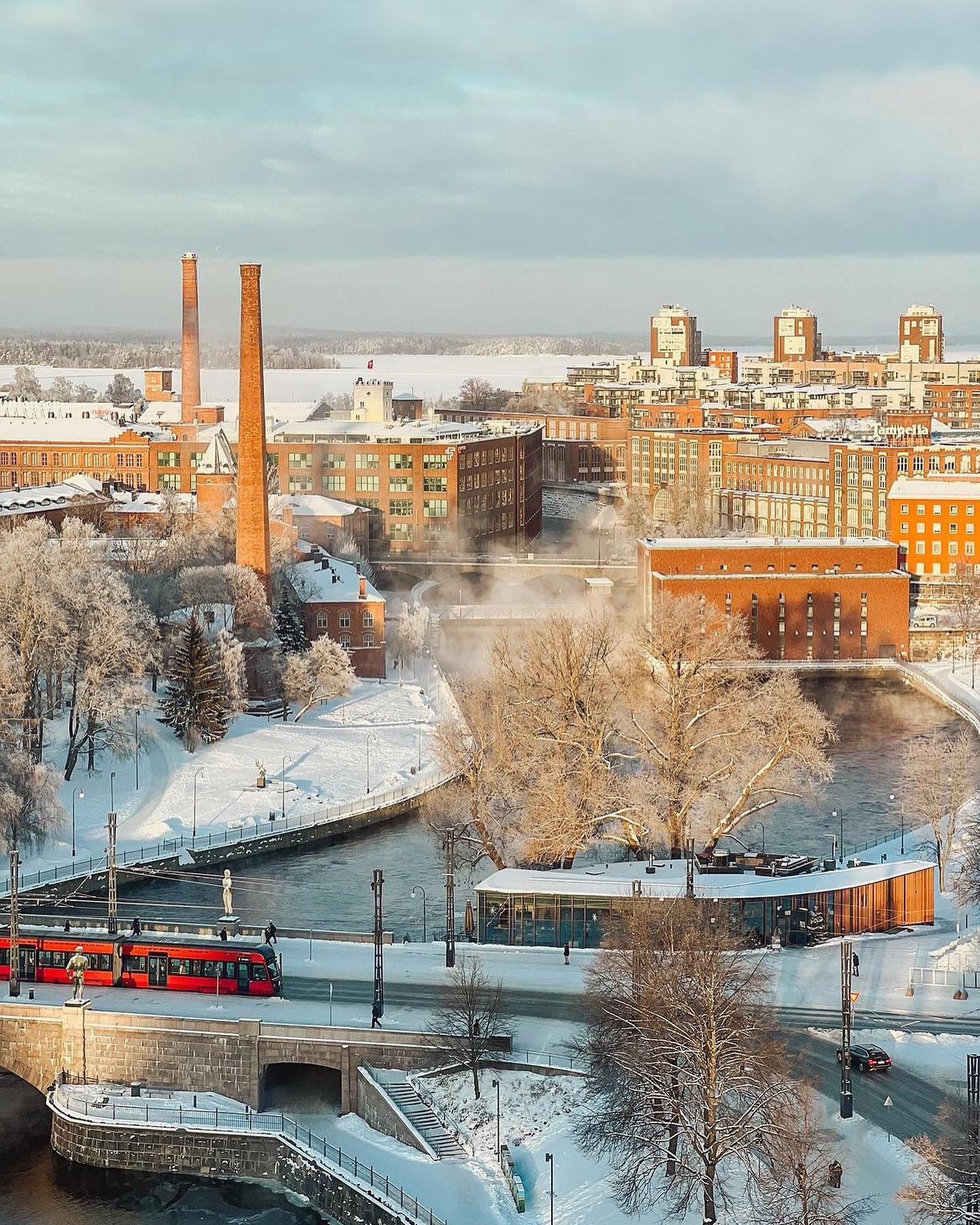 A snowy cityscape featuring a river and industrial buildings with tall chimneys, along the shores of the Tammerkoski rapids. Residential buildings stand in the background, while a red tram crosses a bridge near Koskipuisto Park in Tampere.