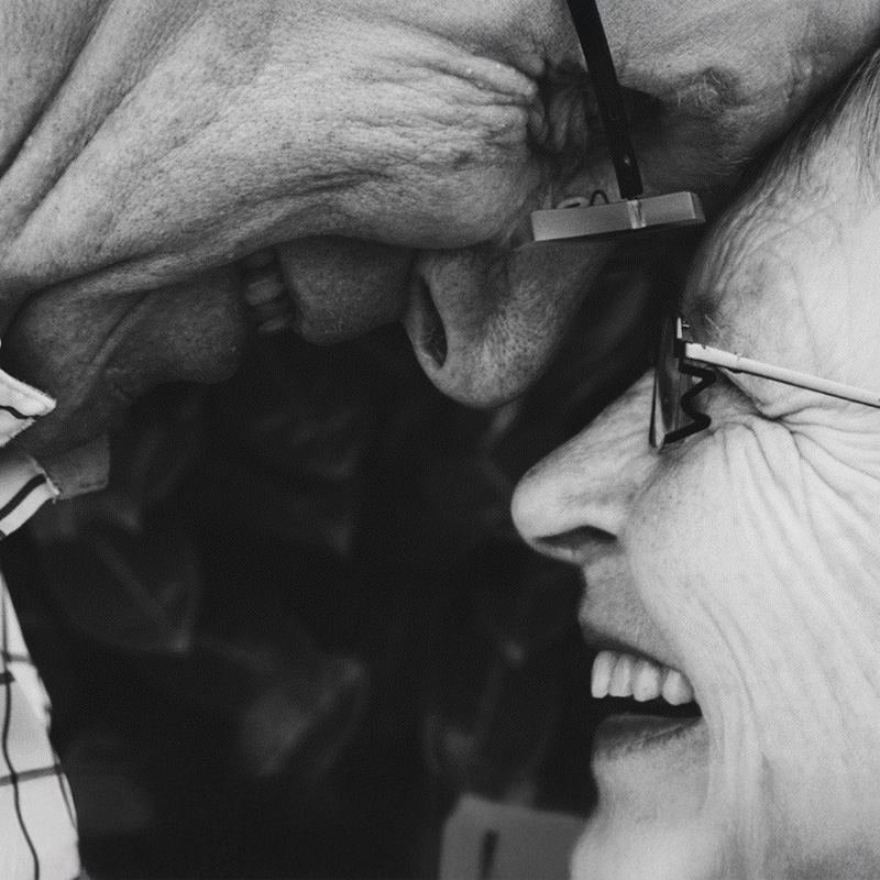 Close-up of two elderly people smiling and touching foreheads, both wearing glasses.