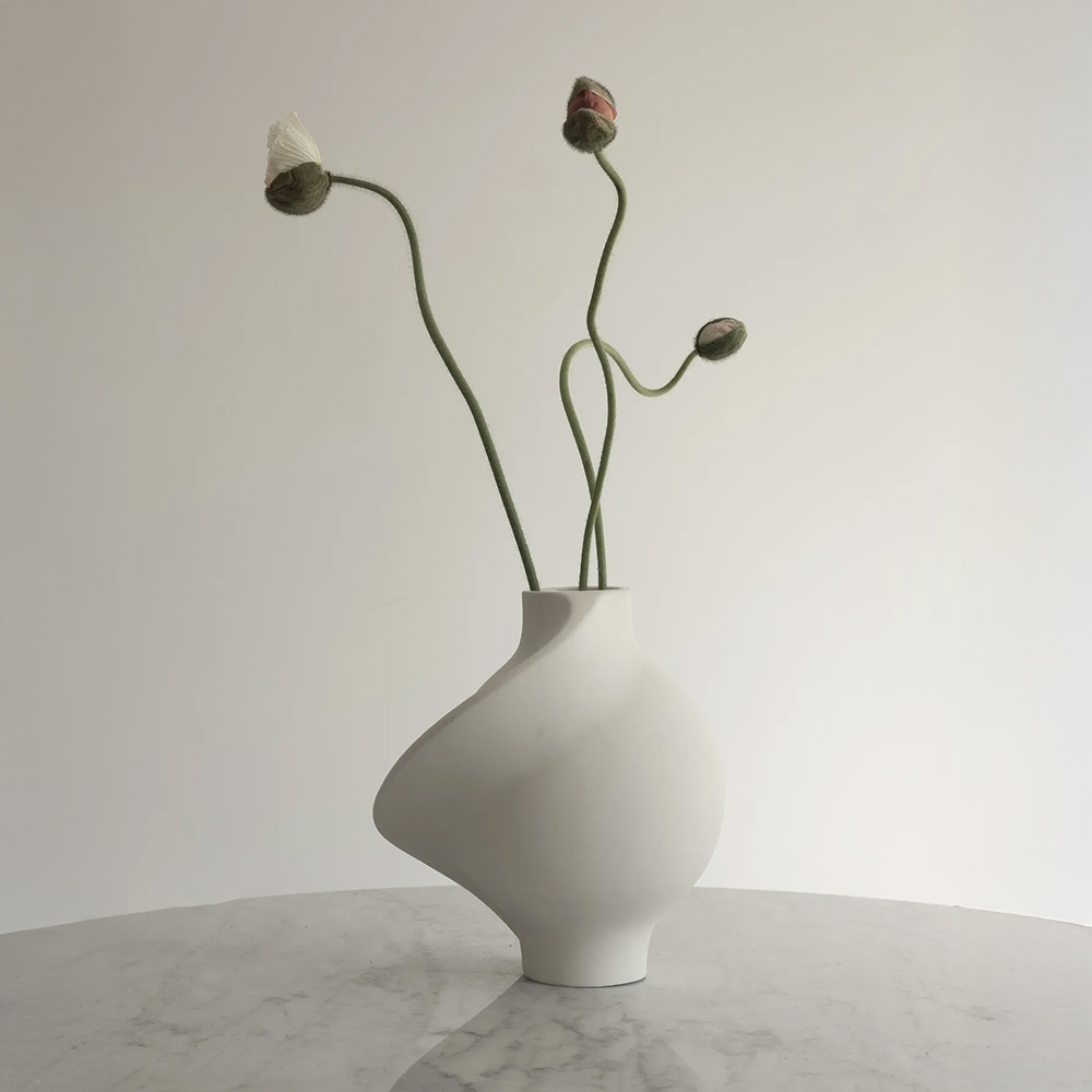 A white, asymmetrical vase with three budding flowers is placed on a marble surface against a plain background.