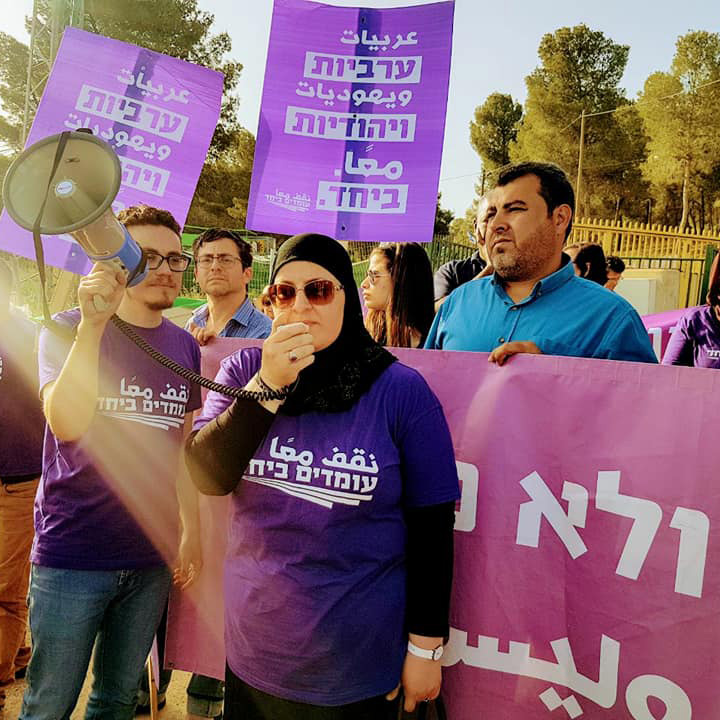 A group of people wearing purple t-shirts hold signs and a megaphone, possibly participating in a protest. There are trees in the background.