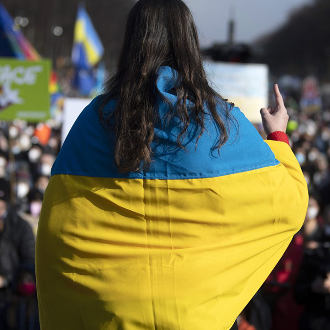 Person draped in a Ukrainian flag gestures with peace sign at a large outdoor gathering.