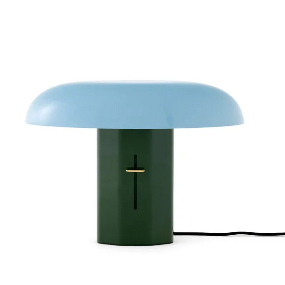 A table lamp with a green cylindrical base and a rounded blue shade, featuring a black vertical slit and a gold accent on the base. A black power cord extends from the lamp.