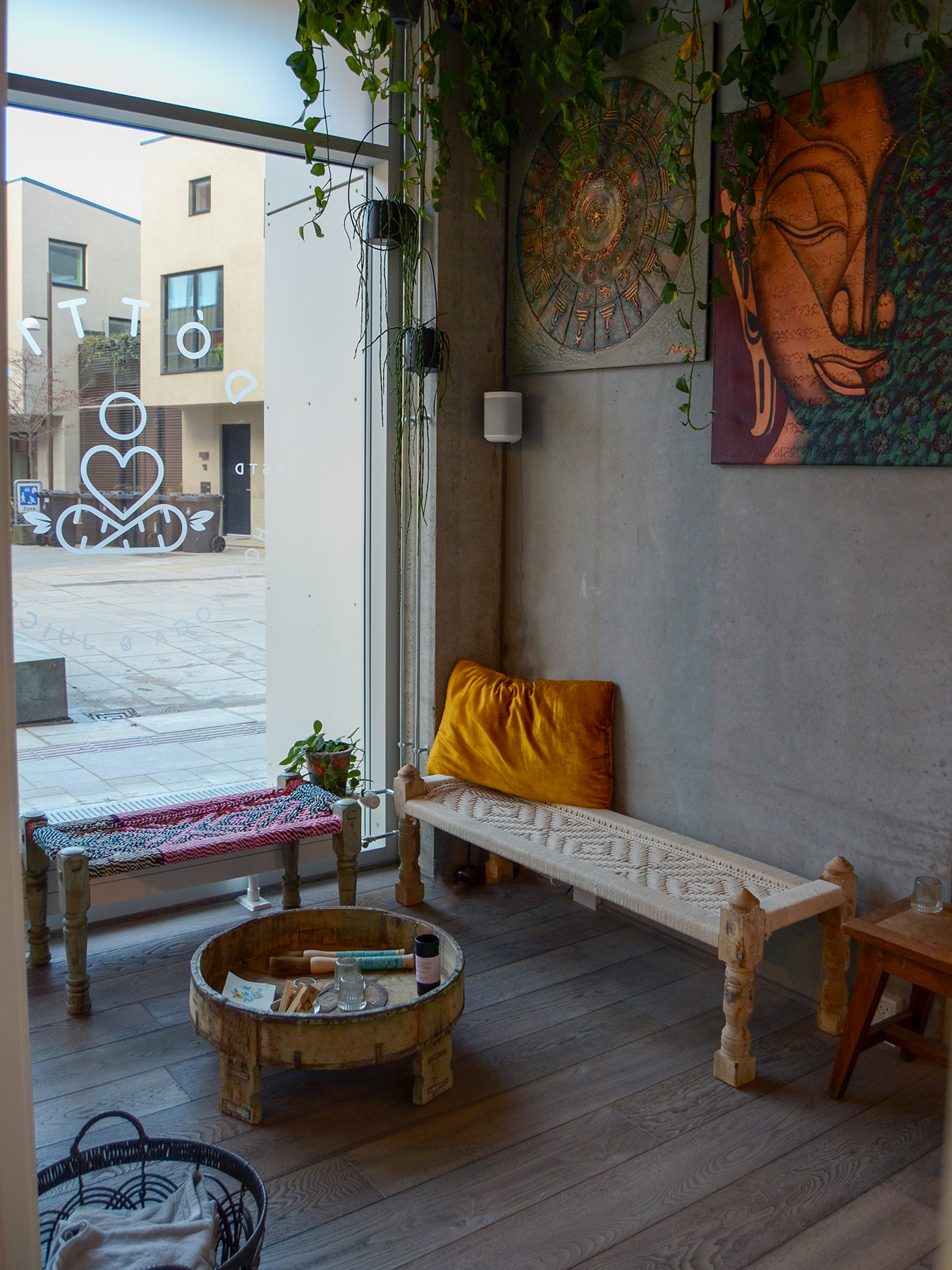 Cozy room with wooden benches, a round coffee table, and wall art. Large window with plants above, brightens the space. Street view in background.