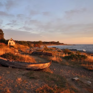 An old wooden boat rests on a grassy shore at sunset, with a small white house in the background and the sea on the right.