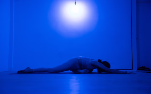 Person stretching on the floor under a blue light, with one leg extended forward and arms reaching ahead.