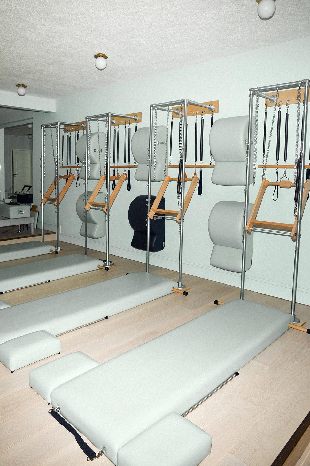 A minimalist Pilates studio with several Cadillac reformer machines lined up against a light-colored wall, each equipped with padded platforms and workout attachments.