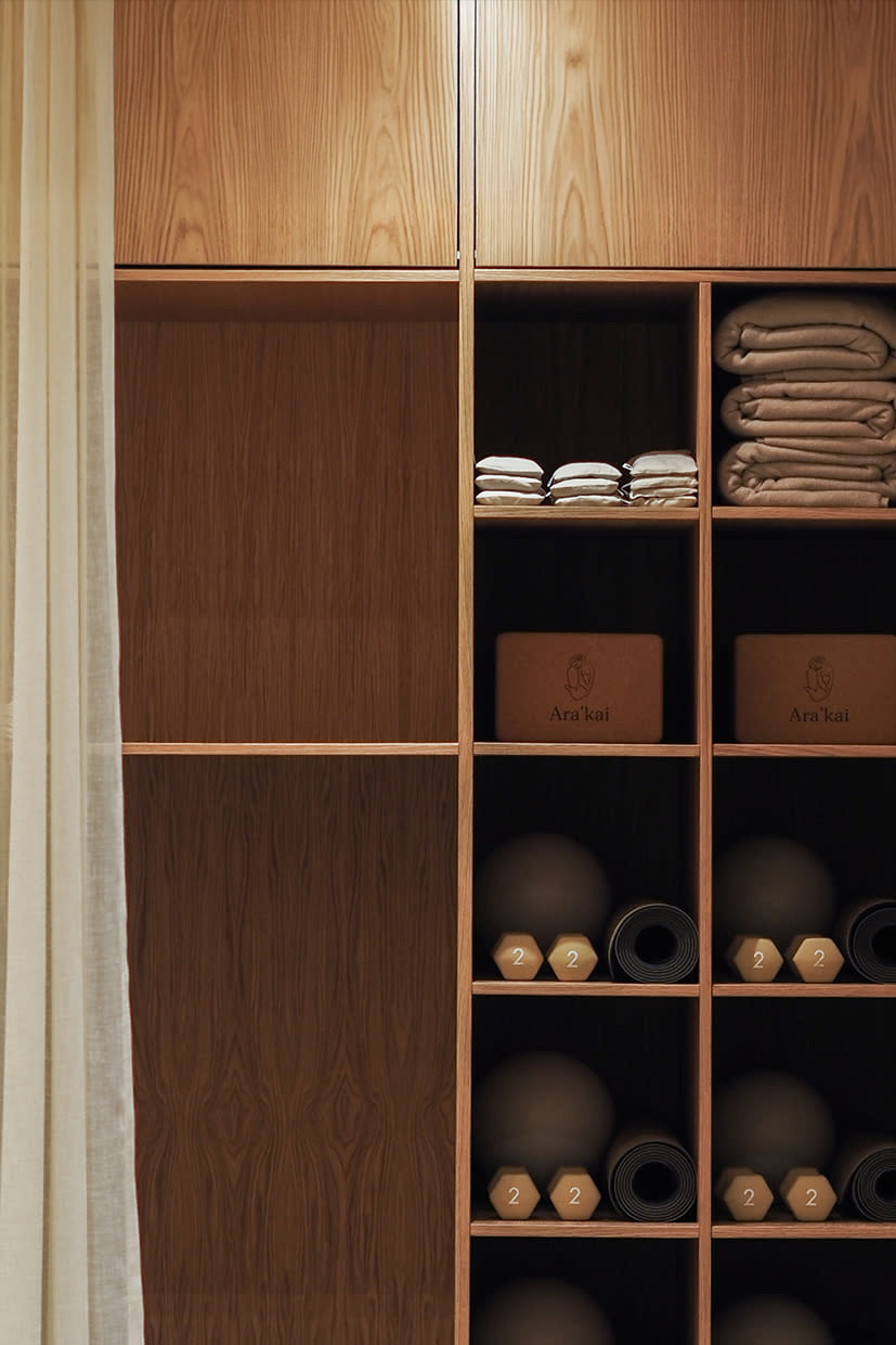 A wooden shelving unit with rolled yoga mats, towels, folded blankets, and boxes, partially covered by a light curtain.