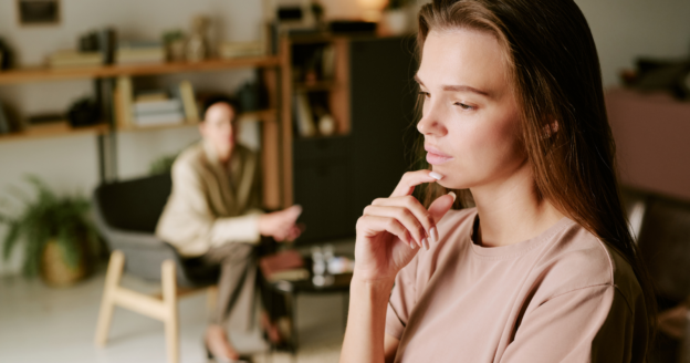 A woman looks thoughtful while sitting in the foreground. In the background, a person sits on a chair, possibly a therapist, in a room with shelves and plants.