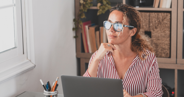 Woman wearing glasses and a striped shirt sits at a desk with a laptop, looking thoughtfully out a window. Shelves with books and plants are in the background.