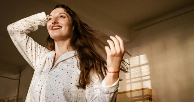 A woman in light-colored pajamas stands indoors, smiling and running her hand through her long hair in soft, natural light.