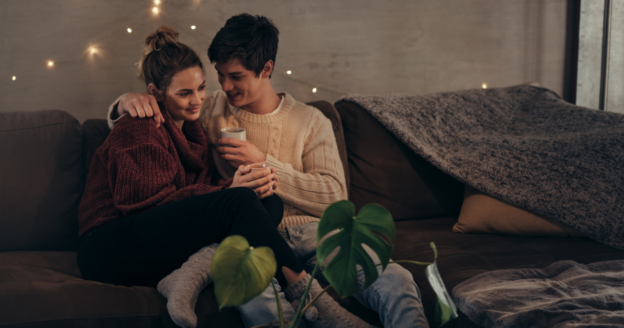 A couple sits close together on a couch, with the man holding a mug. Cozy blankets and string lights are in the background, and a green plant is in the foreground.