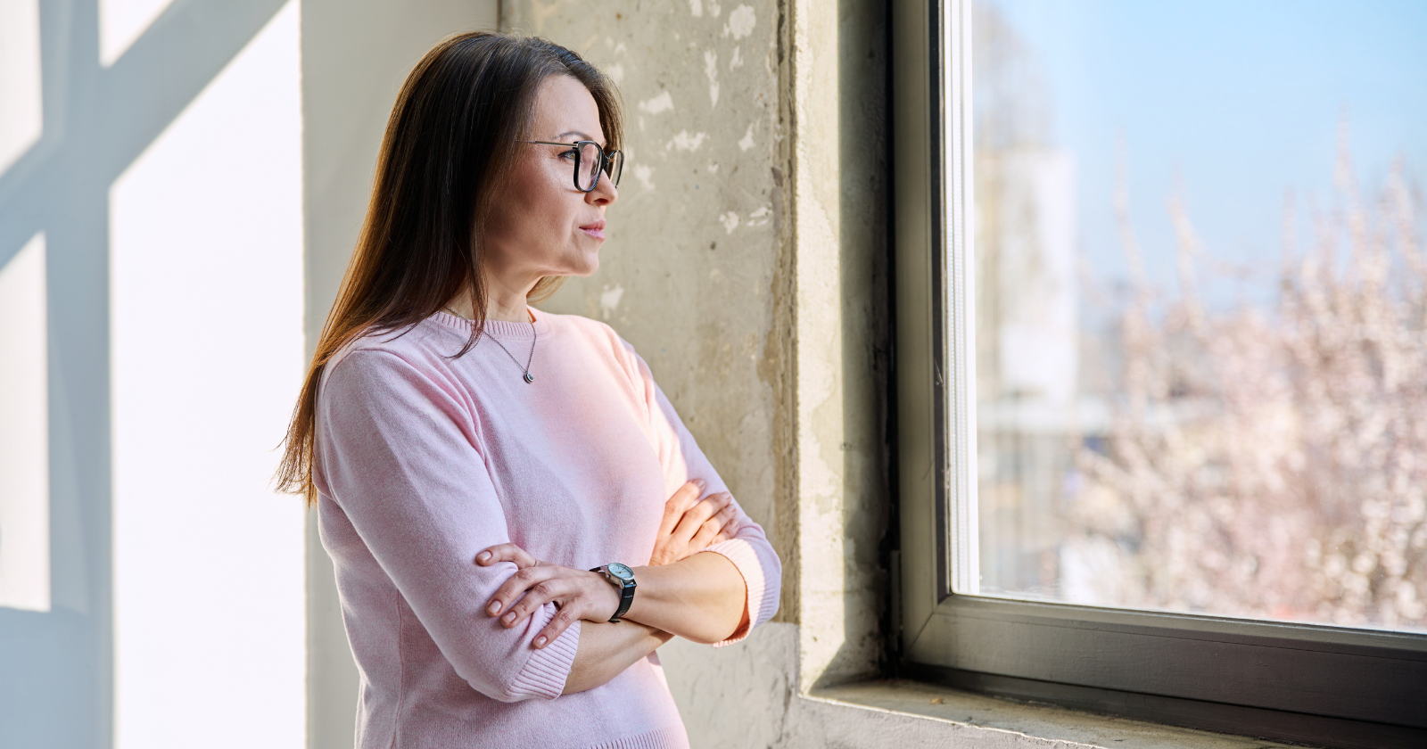 Woman with long hair and glasses, wearing a pink sweater, stands indoors with arms crossed, looking out of a window at a bright, sunlit scene.