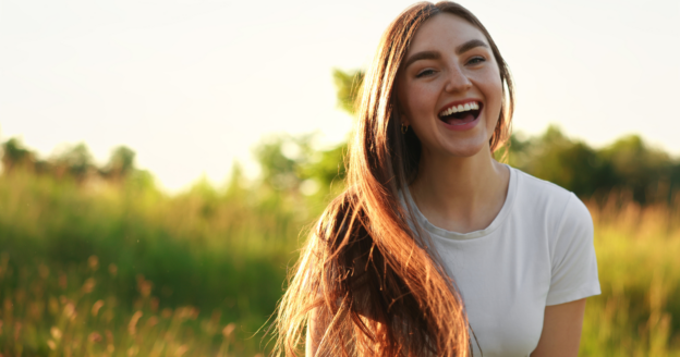 A young woman with long brown hair and a white t-shirt smiles outdoors in a sunlit grassy field.
