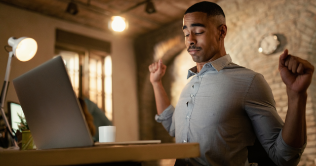 A man in a button-up shirt stretches at his desk in front of an open laptop, with a coffee mug nearby and warm lighting in the workspace.