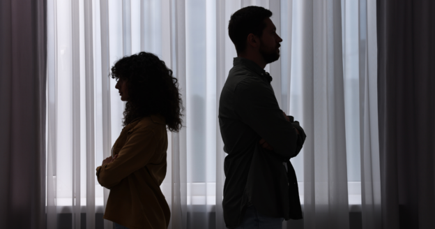 Two people standing back to back with arms crossed in front of a window with sheer curtains, both appearing distant and not facing each other.