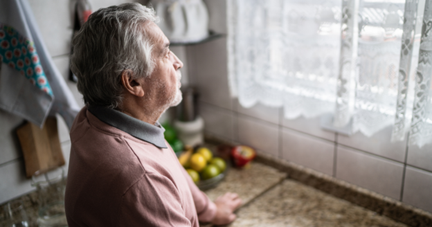 An older man with gray hair stands in a kitchen, looking out a window with lace curtains; fruit is on the counter beside him.