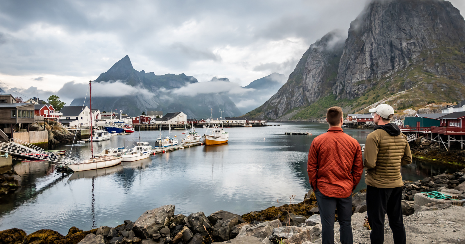 Two people stand on rocks overlooking a harbor with boats, traditional houses, and steep mountains in the background under cloudy skies.