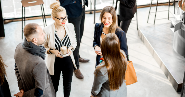 A group of professionally dressed people stand in a circle, conversing and smiling at an indoor networking event. One woman holds a cup of coffee.