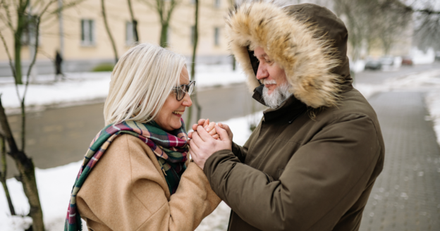 Two older adults stand outside on a snowy day, holding hands and facing each other, both dressed in winter coats and scarves.