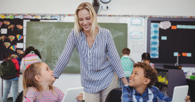 A teacher stands between two smiling students at their desks in a classroom; other children are visible in the background near a chalkboard.