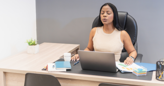 A woman sits at a desk with a laptop, eyes closed and lips pursed, appearing to take a deep breath. Office supplies and a coffee cup are on the desk.
