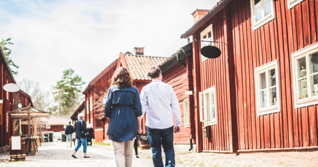 Two people walk down a cobblestone street lined with red wooden buildings on a sunny day.
