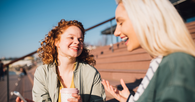 Two women sit on outdoor steps, talking and smiling. One holds a reusable cup while the other gestures with her hand. The sky is clear and sunny.