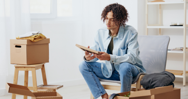 A person in a denim jacket sits in a chair holding a framed photo, surrounded by cardboard moving boxes and packed belongings in a bright room.