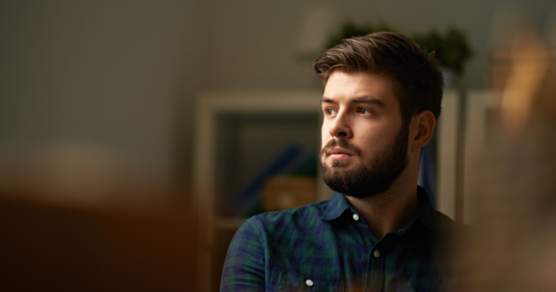 A man with a beard and short hair, wearing a dark plaid shirt, looks thoughtfully to the side in a softly lit indoor setting.