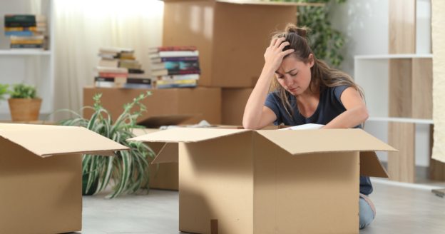 A woman sits on the floor surrounded by cardboard boxes, looking stressed with her hand on her head, in a room with unpacked belongings.