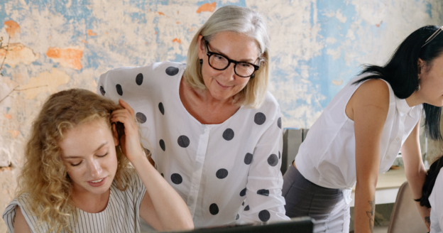Three women collaborate at a desk, with one standing and assisting another who is seated at a computer, in a casual office setting with a textured wall in the background.