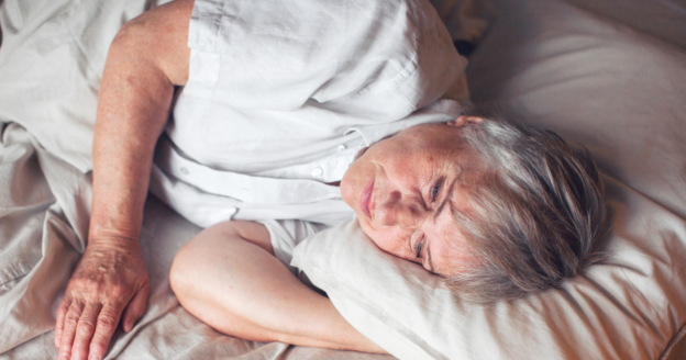 Elderly person with gray hair lying on their side in bed, resting their head on a pillow with a neutral expression.