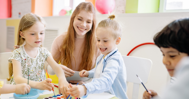 A teacher and three young children sit at a table, smiling and painting together in a brightly lit classroom.