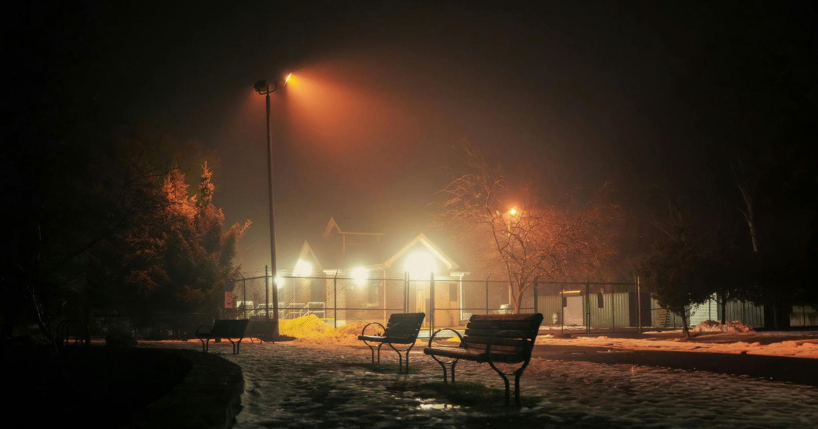A dimly lit park at night with two empty benches, wet ground, scattered snow, and a house illuminated in the background under streetlights.