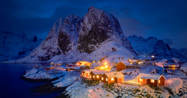 Snow-covered red cabins with glowing lights sit by the water at night, surrounded by mountains in a winter landscape.