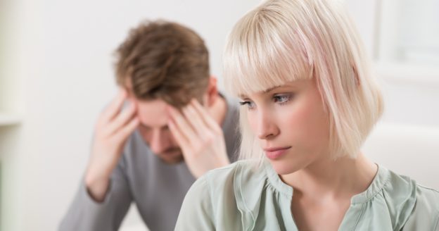 A woman looks away with a serious expression while a man sits behind her, holding his head in his hands, appearing stressed.