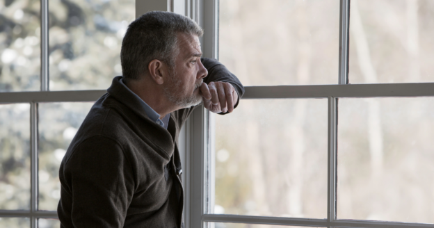 A middle-aged man with gray hair and a beard stands indoors, leaning on a window frame and looking outside with a thoughtful expression.