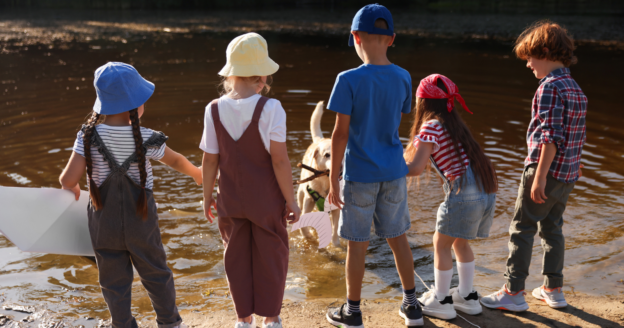Five children stand at the edge of a shallow pond, facing away, watching a dog in the water on a sunny day.