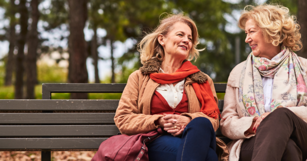 Two older women sit on a park bench, facing each other and talking, with trees and greenery in the background.