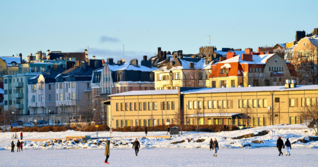 People walk on snow-covered ground in front of colorful buildings under clear blue sky in a winter cityscape.