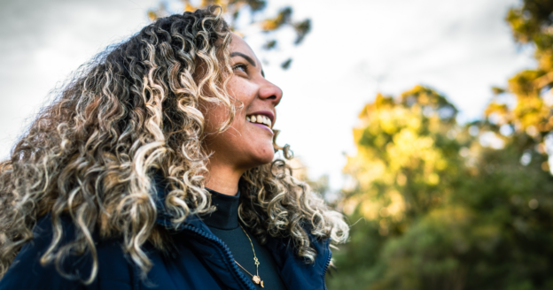 A woman with curly, highlighted hair smiles while looking to the side outdoors, with trees and greenery in the background.