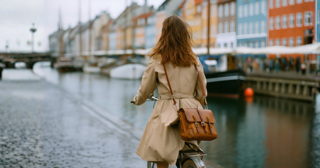 A person in a beige coat rides a bicycle along a canal lined with colorful buildings and boats on an overcast day.