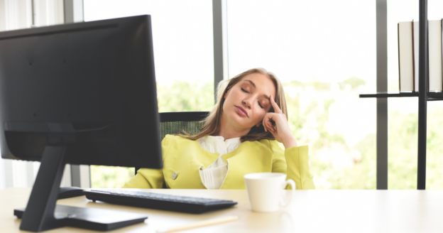 A woman in a yellow blazer sits at a desk with a computer, resting her head on her hand and appearing to be asleep. A coffee cup and papers are also on the desk.